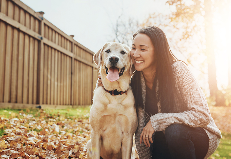 Woman and dog sitting together in a backyard with wooden fence and trees in the background.