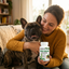 Woman holding a dog and a bottle of PYWGANIX YeastGuard supplement in a home setting.